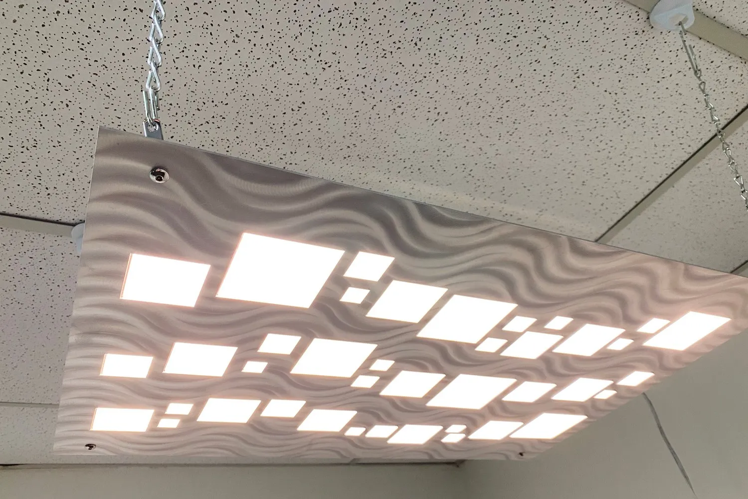 Suspended ceiling luminaire with rows of glowing CoreGLO rectangles set into a wave-textured metal face