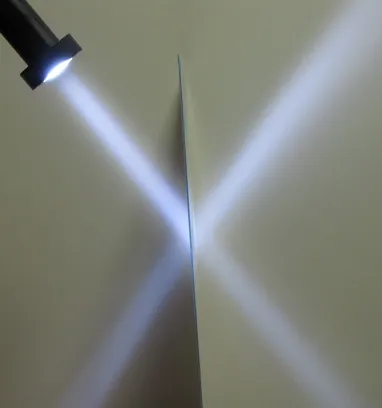 Flashlight beam hitting an edge-on sheet of Daylighting Fabric®: one part of the beam passes through and another part is redirected, forming an X of light on the wall