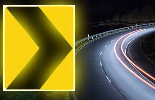 Yellow chevron road sign glowing at night beside a curving road