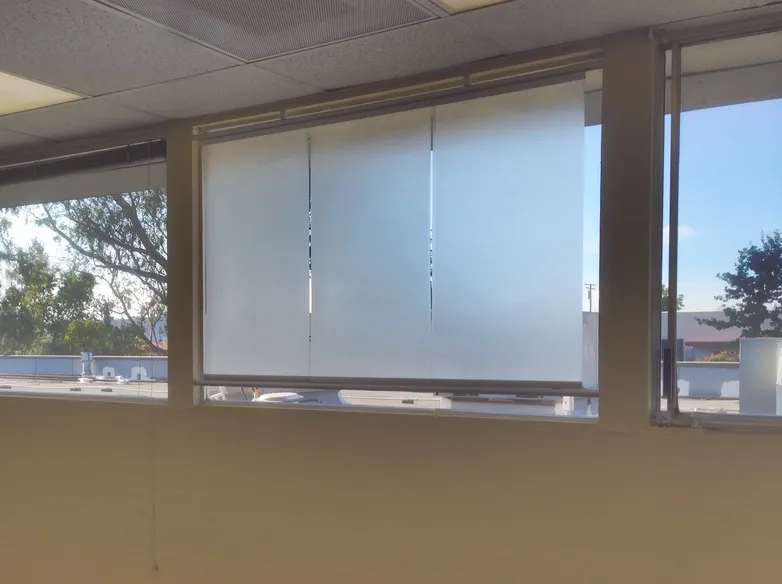 Office window fitted with three translucent Luminyl™ shade panels, with trees and rooftops visible through the clear windows beside it