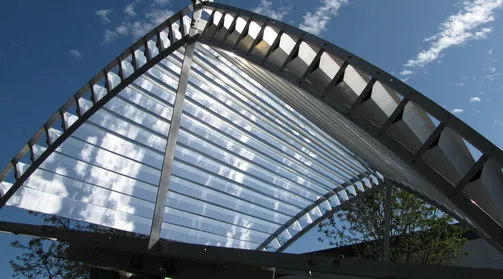 Arched slat-array concentrator seen from below against a blue sky: an open frame holding rows of parallel reflective slats